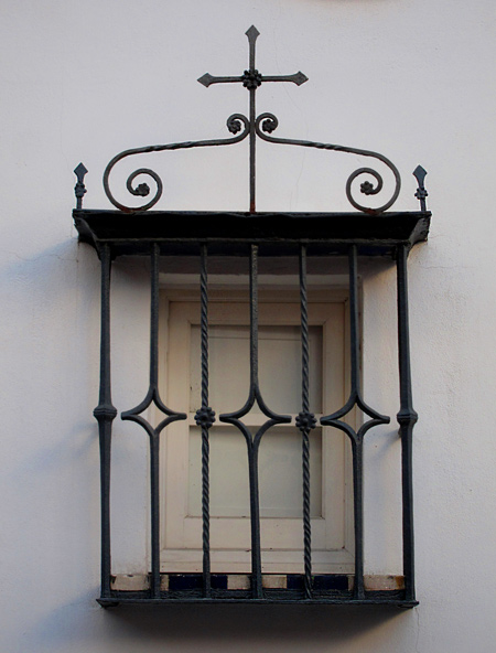 A historic black wrought iron window grille with cross and black and white decorative tile shelf. A photo taken on a historic street in Seville, Spain near the Royal Alcazar Palace