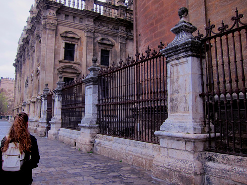 a girl with a backpack walks past the historic walls, columns and wrought iron fence details outside Catedral de Sevilla in Seville, Spain