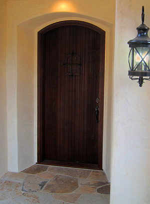 A rustic Spanish style arch top entry door withspeakeasy iron grille and a flagstone step at a Montecito California cottage